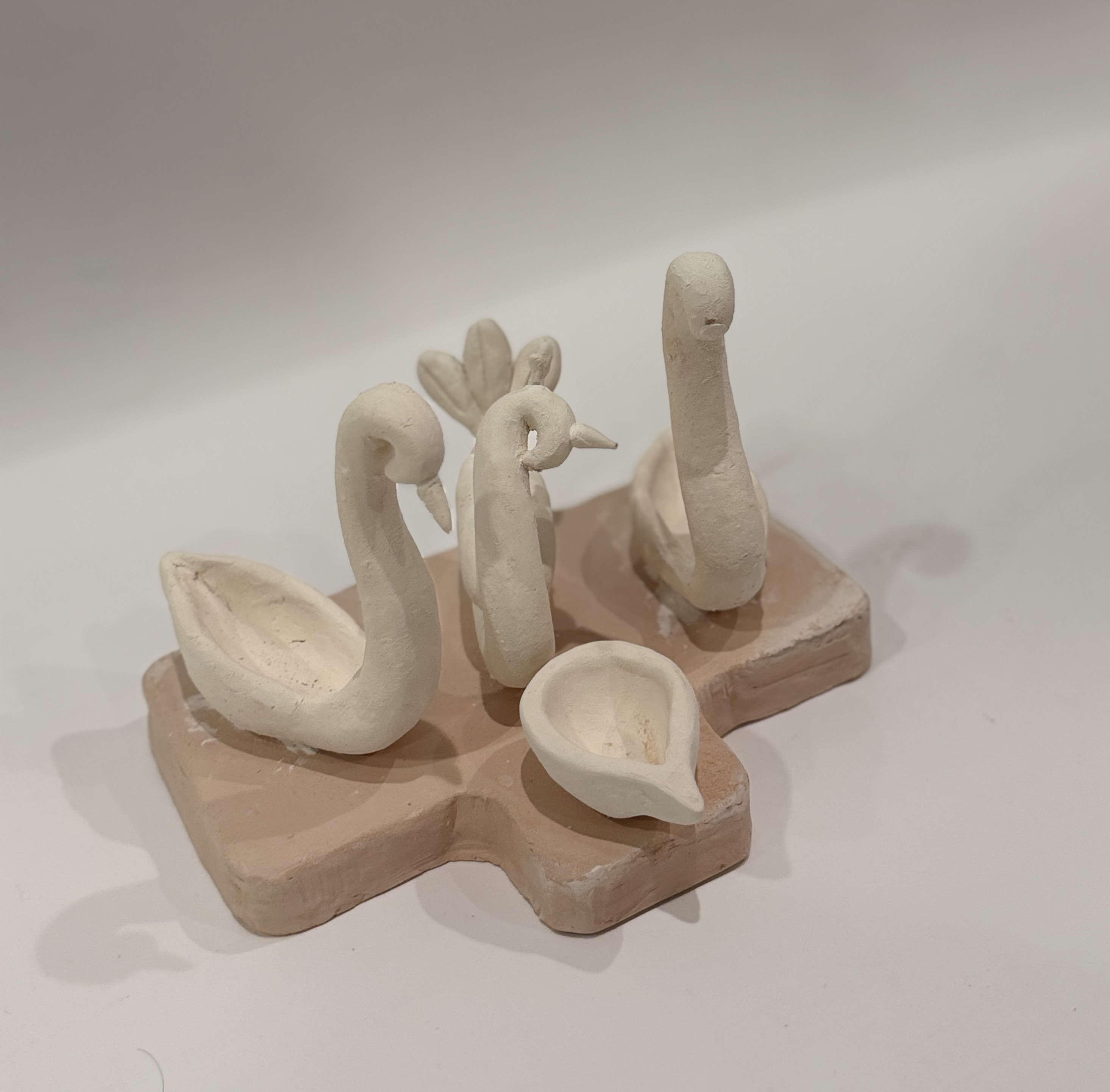 An unfired sculpture of swans arranged around oil lamps