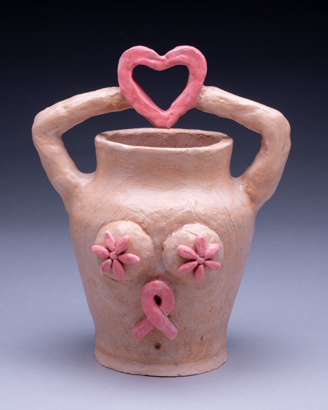 A hand-built vessel with raised arms forming a heart and a pink ribbon on its face